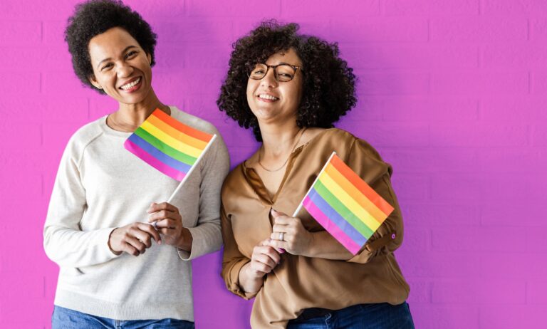 This is an image of two queer women holding the Pride flag on a pink wall.