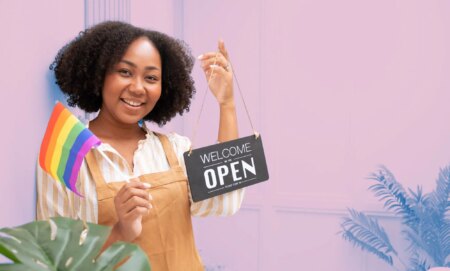 This is an image of a Black woman holding an "we're open" sign in one hand and a Pride flag in the owner. She is wearing a brown apron.