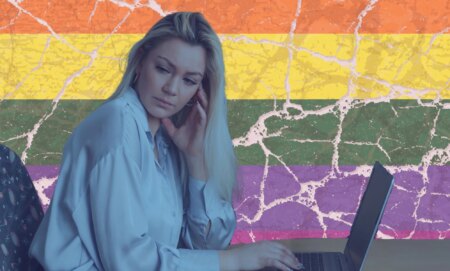 this is a stylized image of a woman looking unhappy or distressed at a computer. She is in a blue tint and behind her is the pride flag, but it is cracked.