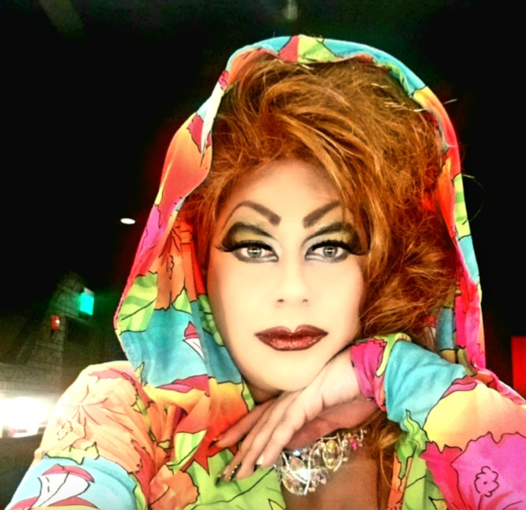 L'image montre la drag queen de Nashville, Veronika Electronika, dans une tenue colorée avec une capuche relevée sur ses cheveux, elle pose sa tête sur sa main et regarde la caméra.