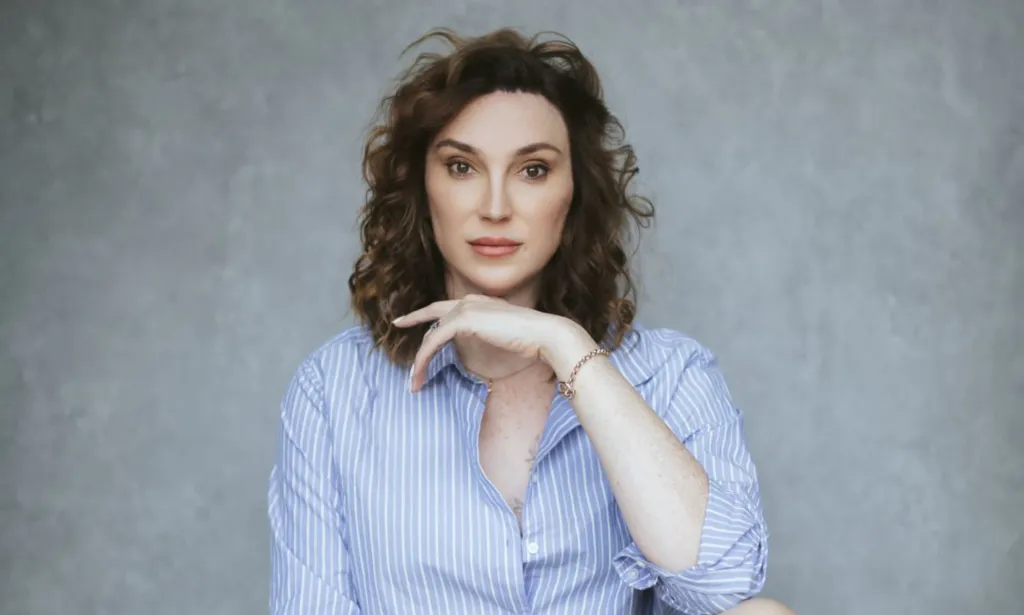 Juno Dawson pose avec sa main sur son menton, vêtue d'une chemise bleue, sur un fond gris.