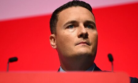 A politician at a podium with two microphones stands infront of a red screen.