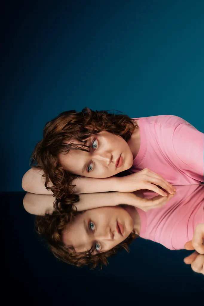 Dee Allum au festival Fringe d'Édimbourg. Dee porte un t-shirt rose posé sur une table en miroir. Son reflet dans le miroir regarde la caméra.