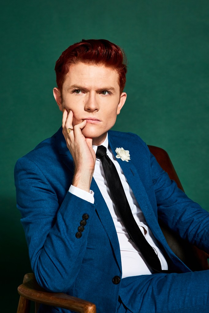 Rhys Nicholson : Félicitations pour cette grande fête ! Ryan porte un costume bleu et est assis sur une chaise marron sur fond vert. Il pose son visage sur sa main, l'air sérieux.