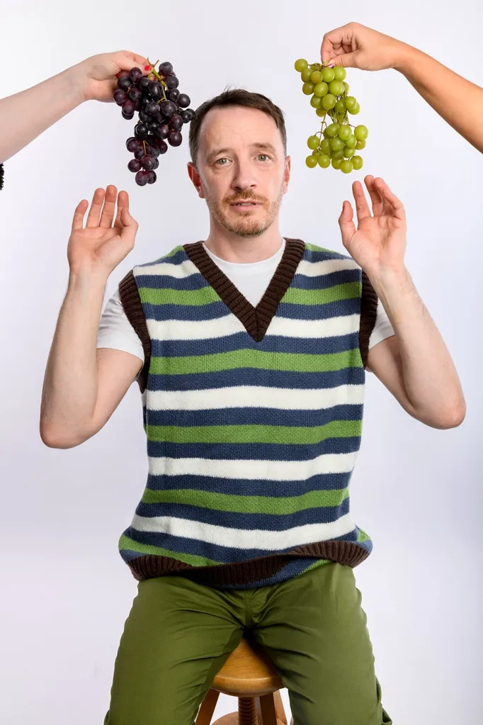 Gearóid Farrelly : Gearóid Rage. Gearóid est assis sur un stand avec des raisins rouges et blancs autour de sa tête. Il porte un gilet rayé blanc, bleu et vert.