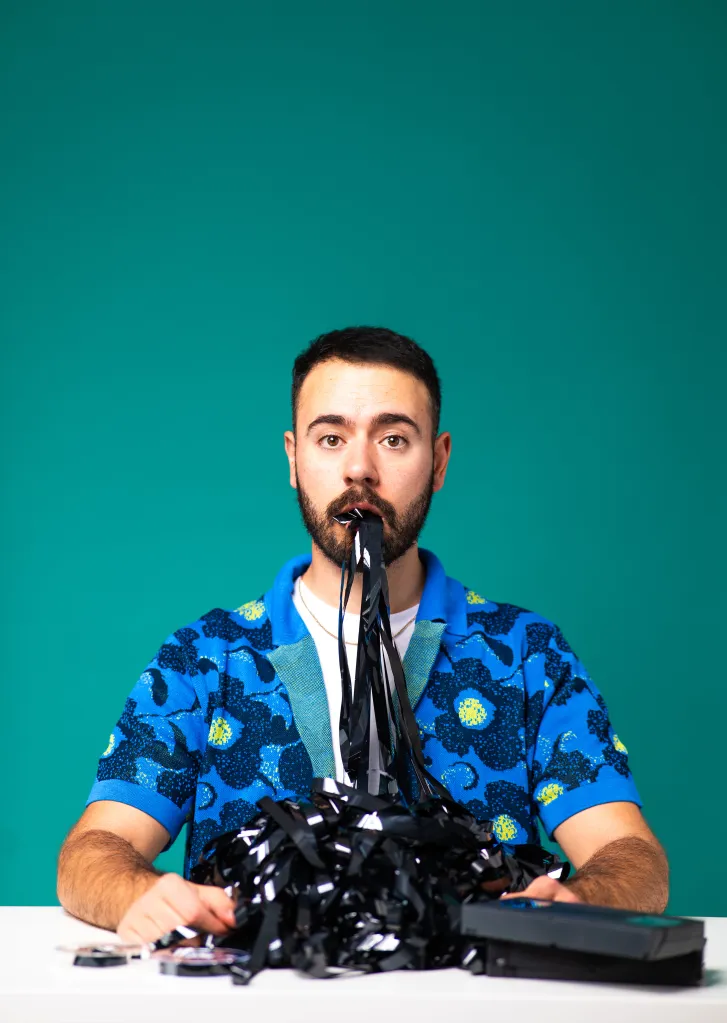Anesti Danelis : Intelligence artificielle. Anesti est assis à un bureau, vêtu d'un haut bleu à fleurs avec une cassette VHS sortant de sa bouche et s'empilant sur la table devant lui.