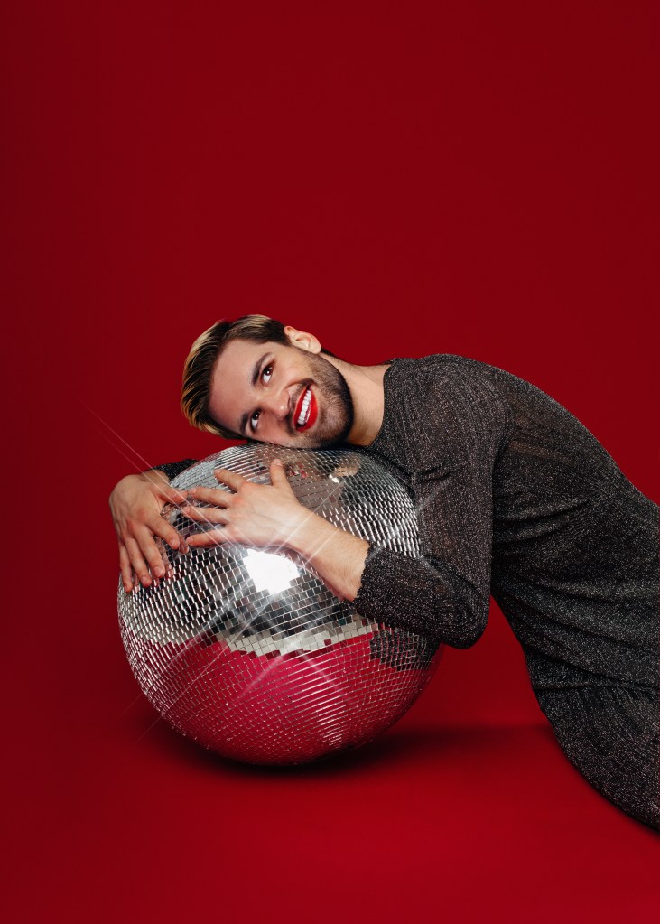 Will Owen dans un look scintillant avec du rouge à lèvres rouge serrant une boule à facettes.