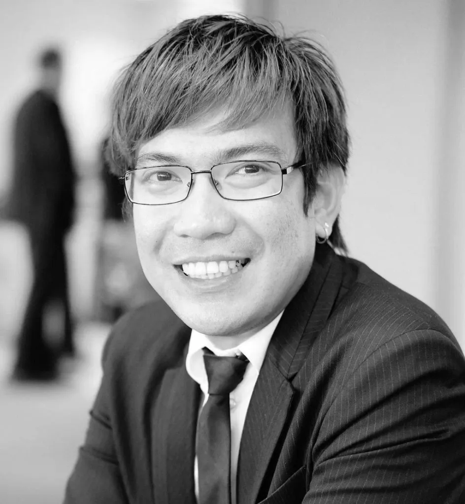 Il s'agit d'un portrait en noir et blanc d'un homme asiatique. Il porte des lunettes à monture métallique et a les cheveux courts et hirsutes. Il porte un costume noir.