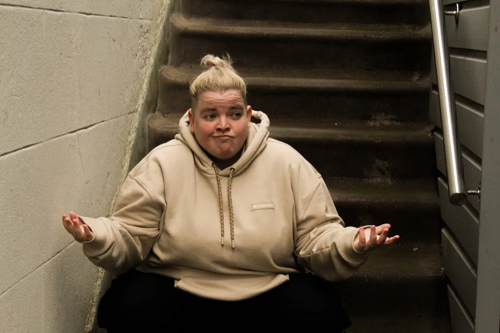 Kim Blythe : Might As Well au festival Fringe d'Édimbourg. Kim porte un sweat à capuche marron clair alors qu'elle est assise au bas d'un escalier.
