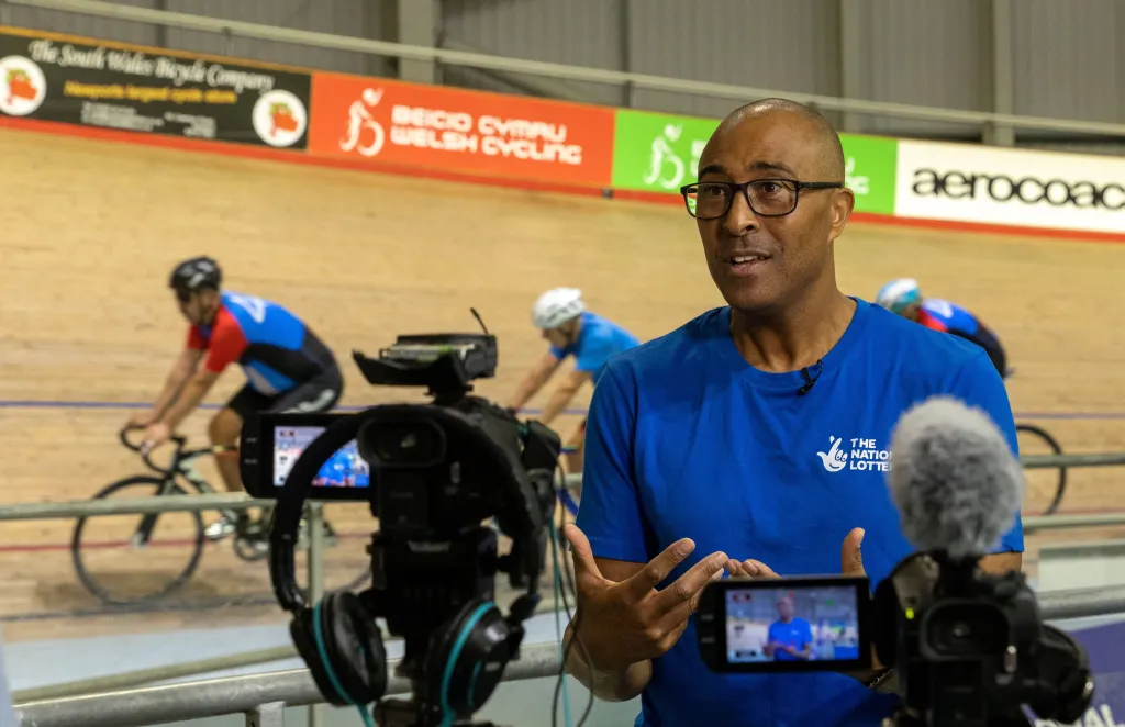 Colin Jackson aux Jeux du Commonwealth de Birmingham 2022, commentant avec la caméra sur lui alors que trois cyclistes passent derrière lui.