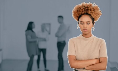 This is an image of a Black woman looking stressed and isolated at work. She has her arms crossed. In the background there are 3 other people blurred.