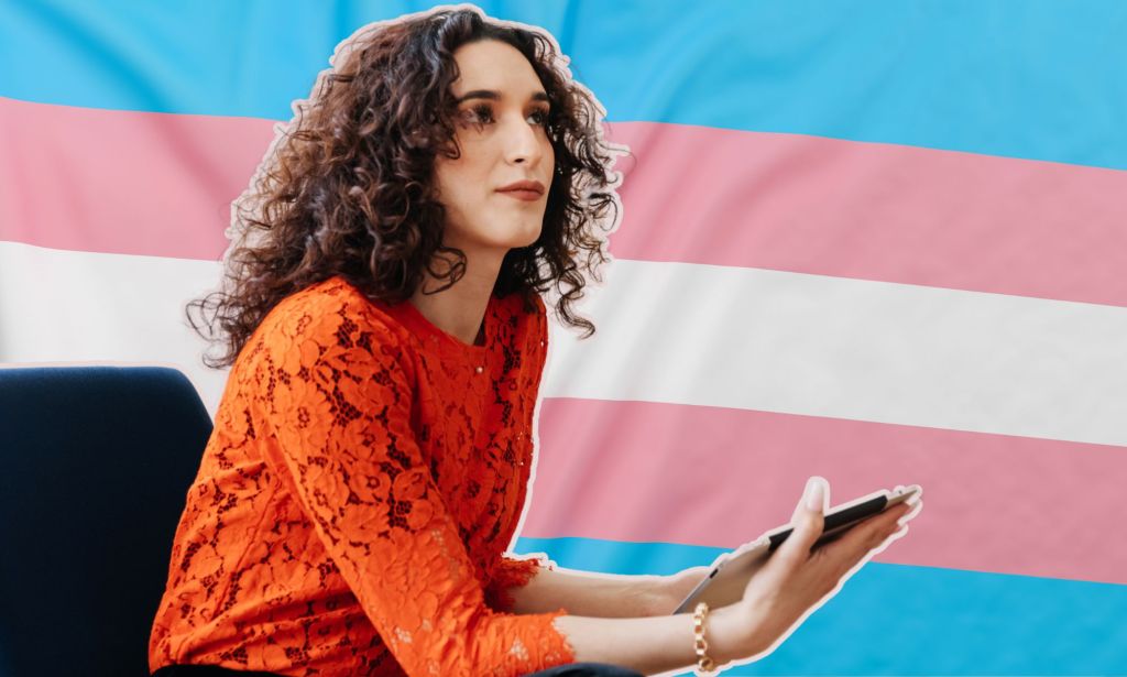 Il s'agit d'une femme transgenre assise sur une chaise. Derrière elle se trouve un graphique du drapeau de la fierté trans. 