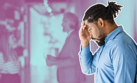 This is an image of a Black man looking frustrated while leaning on a wall. He has short dread locks tied up behind his head. He has a beard and is wearing a blue shirt.