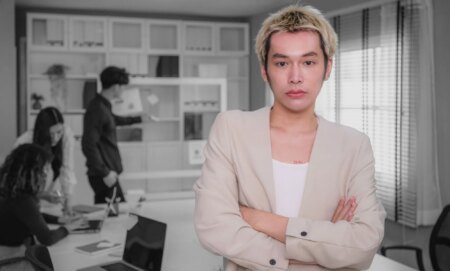 Portrait of young Asian people Non-Binary person standing with arms crossed and colleagues working together in background while discussing on new project at modern office, Corporate business meeting and Successful partners teamwork, Employees engagement and white-collar worker, Finance and Economy, Global Business and Strategy, Looking at camera