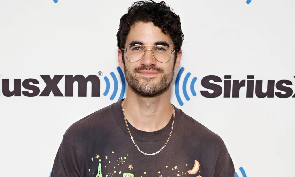 Darren Criss, un acteur non LGBTQ+ devenu célèbre pour ses rôles queer, porte une chemise marron, un collier et des lunettes alors qu'il sourit à la caméra