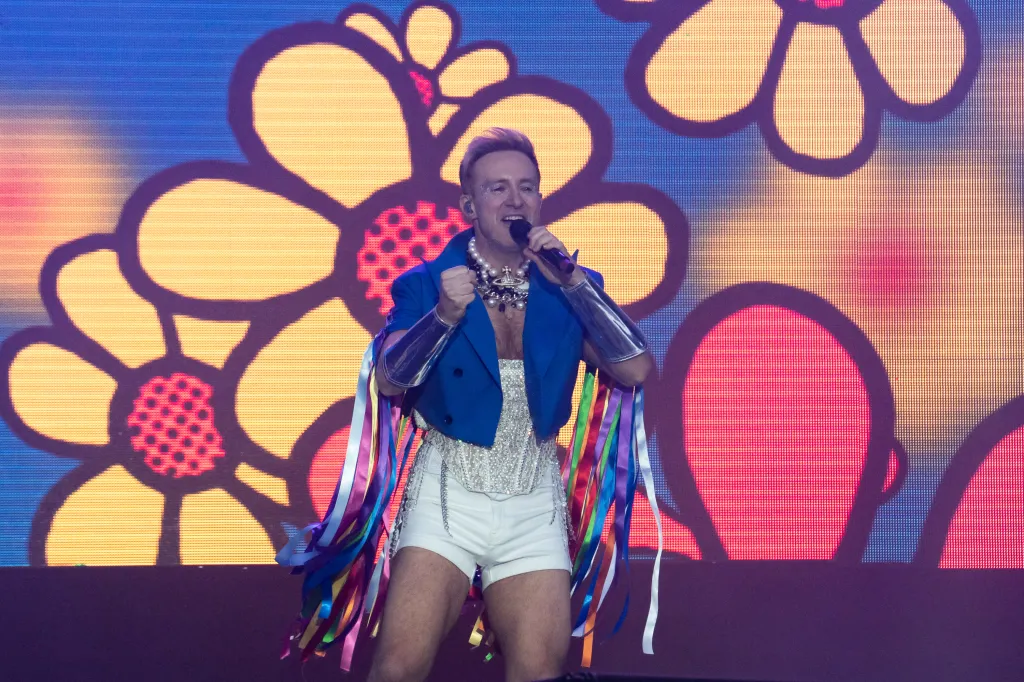 Ian "H" Watkins of Steps se produit lors de la Brighton and Hove 2023 Pride. H joue avec de grandes fleurs de dessins animés derrière lui. Il porte un corset blanc argenté et un short avec un gilet bleu décoré de banderoles aux couleurs du drapeau de la Fierté.