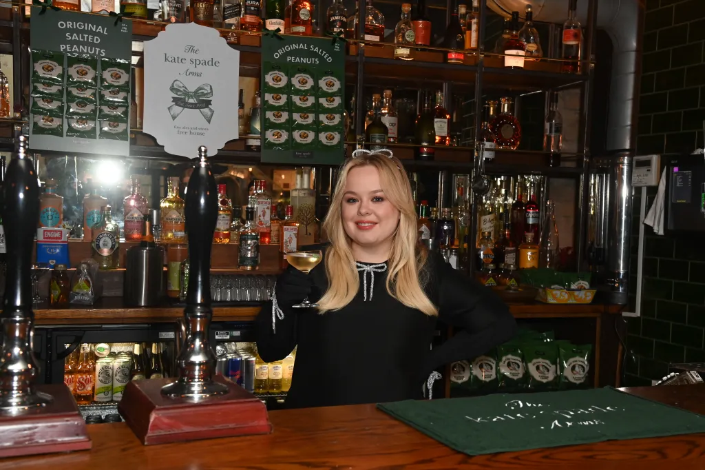 Nicola Coughlan assiste au Kate Spade Arms, elle est debout dans une robe noire derrière le bar avec un verre de vin blanc