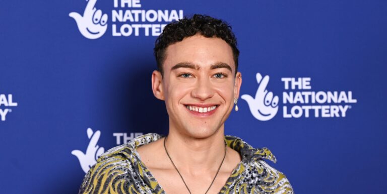 Olly Alexander in a multi-coloured shirt smiling against a blue and white background.