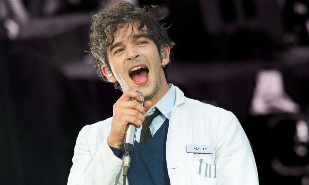Matty Healy du 1975 se produisant sur scène avec une blouse de laboratoire, un pull et une chemise et une cravate.