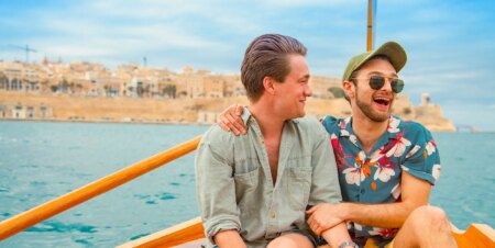 Two men, one in short sleeve colourful t-shirt and the other in a long-sleeve mint shirt, have their arms around each other and are smiling. They are sat on a boat out to sea in Malta.