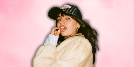 Rebecca Black in a white faux fur jacket and baseball cap, posing with her mouth open and finger and thumb near her face.