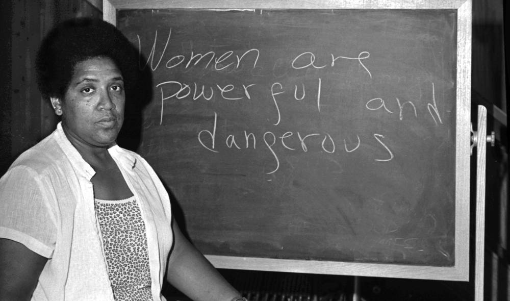 Audrey Lorde standing in front of a chalkboard with the message