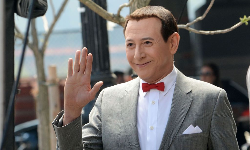 L'acteur Herman Pee-Wee Paul Reubens portant un costume gris, une chemise blanche et un nœud papillon rouge. Il sourit et agit la caméra.