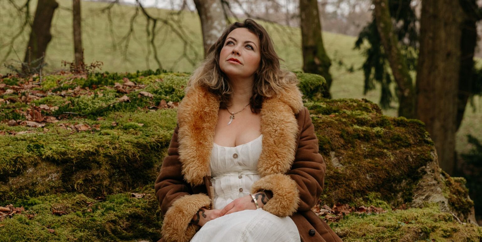 Charlotte Church looking pensive while perching in a forest in a white dress and brown fluffy coat.