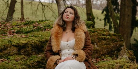Charlotte Church looking pensive while perching in a forest in a white dress and brown fluffy coat.