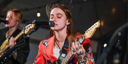 Julien Baker singing into a mic with her eyes closed and her brown hair billowing in the wind above her head. She is holding a guitar and wearing a red blazer and white top.
