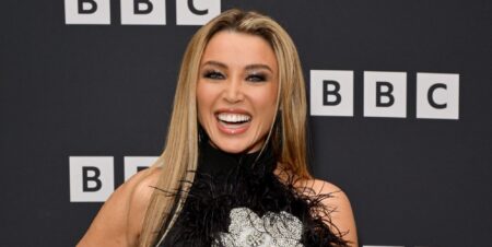 Dannii Minogue smiles as she stands against a BBC branded black background while wearing a black dress.