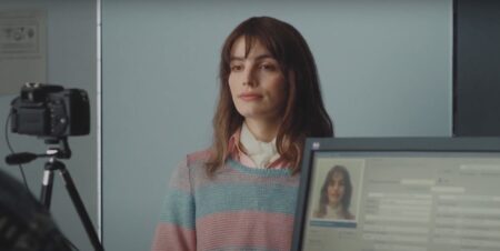Lux Pascal in a blue and pink striped jumped in a scene in new film Miss Carbon. She is stood in front of a blue wall and behind a computer screen and camera.