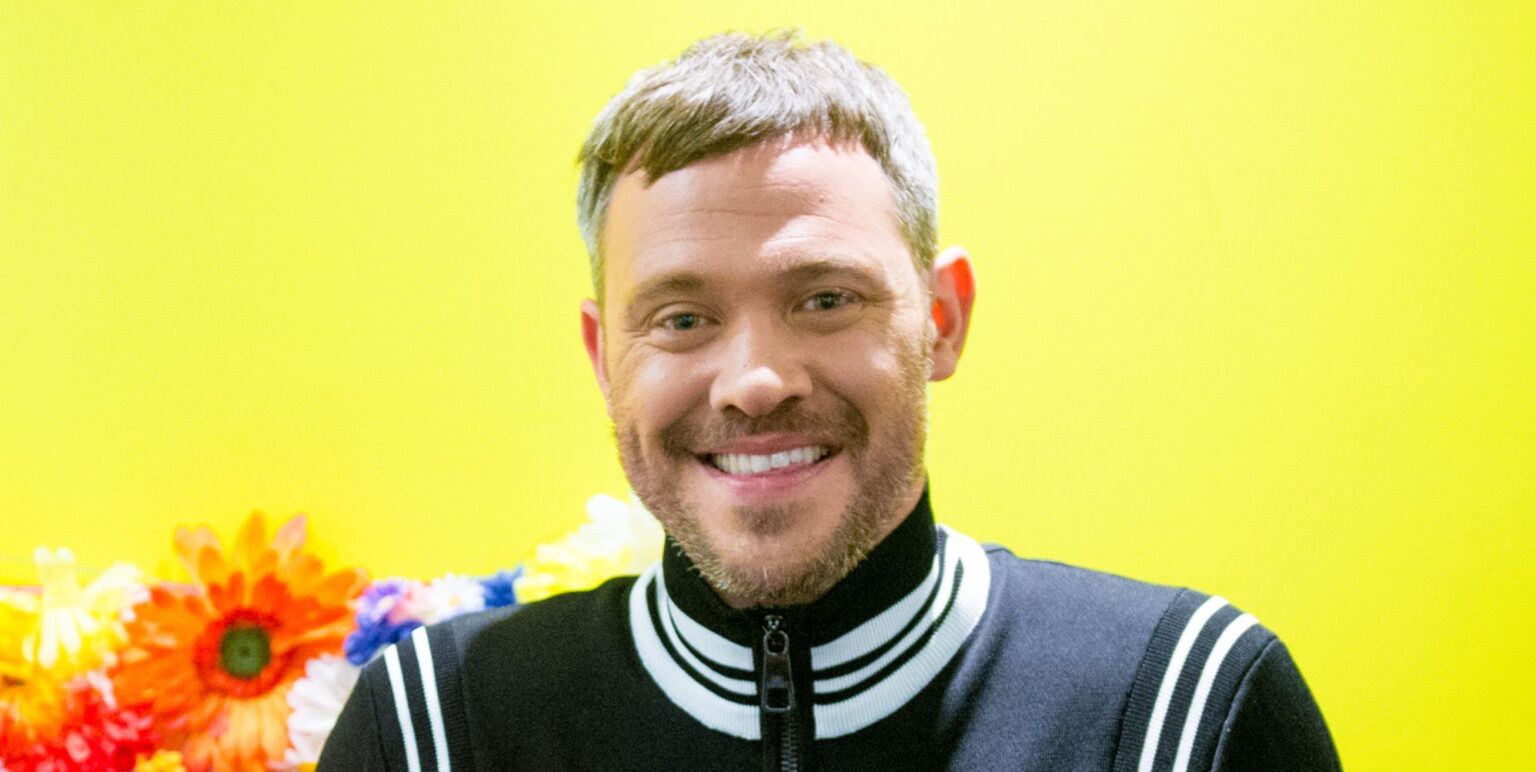 Will Young in a black and white jacket smiling while posing against a yellow and orange background.