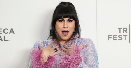 Roz Hernandez a l'air choquée alors qu'elle se tient sur un fond blanc portant une robe violette scintillante avec des poignets moelleux roses.