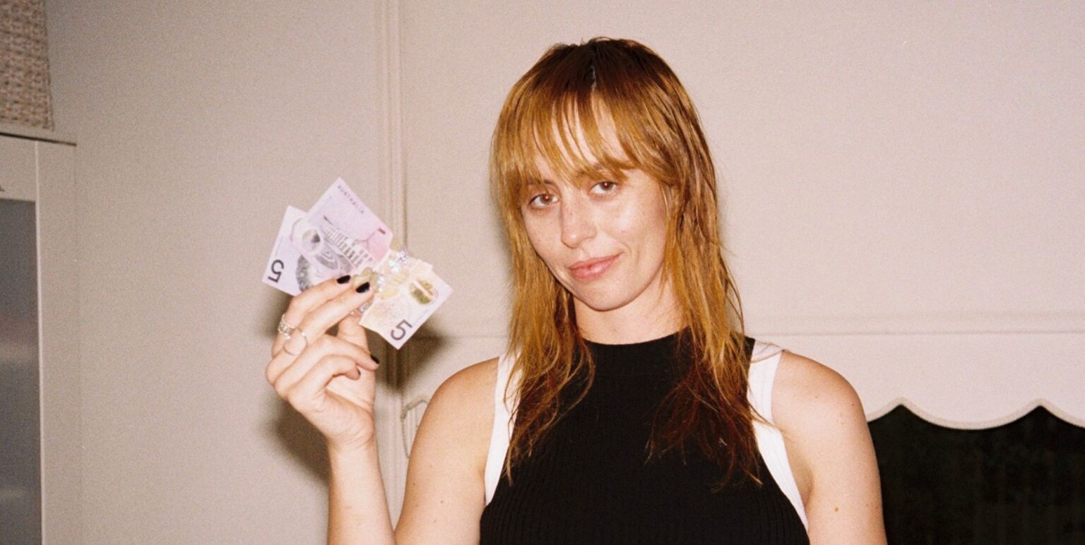 Gordi in a black vest top holding up notes of cash and smiling.