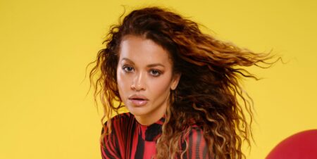 Rita Ora in a black and red top posing against a yellow and red background.