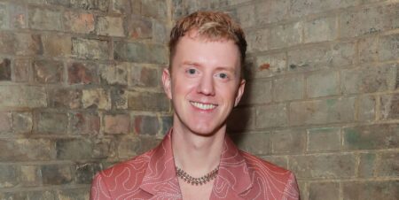 James Bar in a peach blazer and necklace smiling while posing against a brick wall.