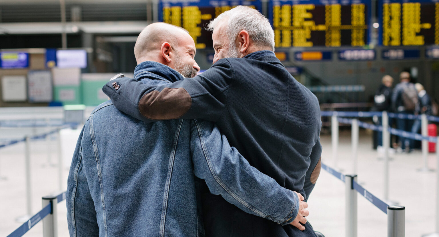 A gay couple walking and embracing each other closely while making their way through an airport, on their way to the check-in before catching a flight together.