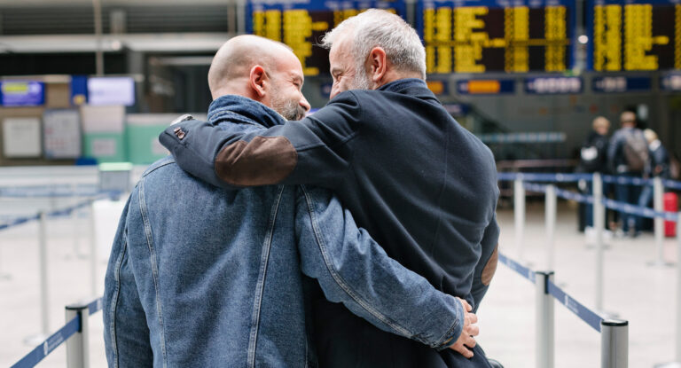 A gay couple walking and embracing each other closely while making their way through an airport, on their way to the check-in before catching a flight together.