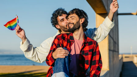 Mid-adult male gay couple embracing while holding LGBTQ+ flags outdoors in Barcelona in Spain.