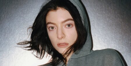 Lorde in a grey hoodie with brown hair poking out while standing against a grey background as she released