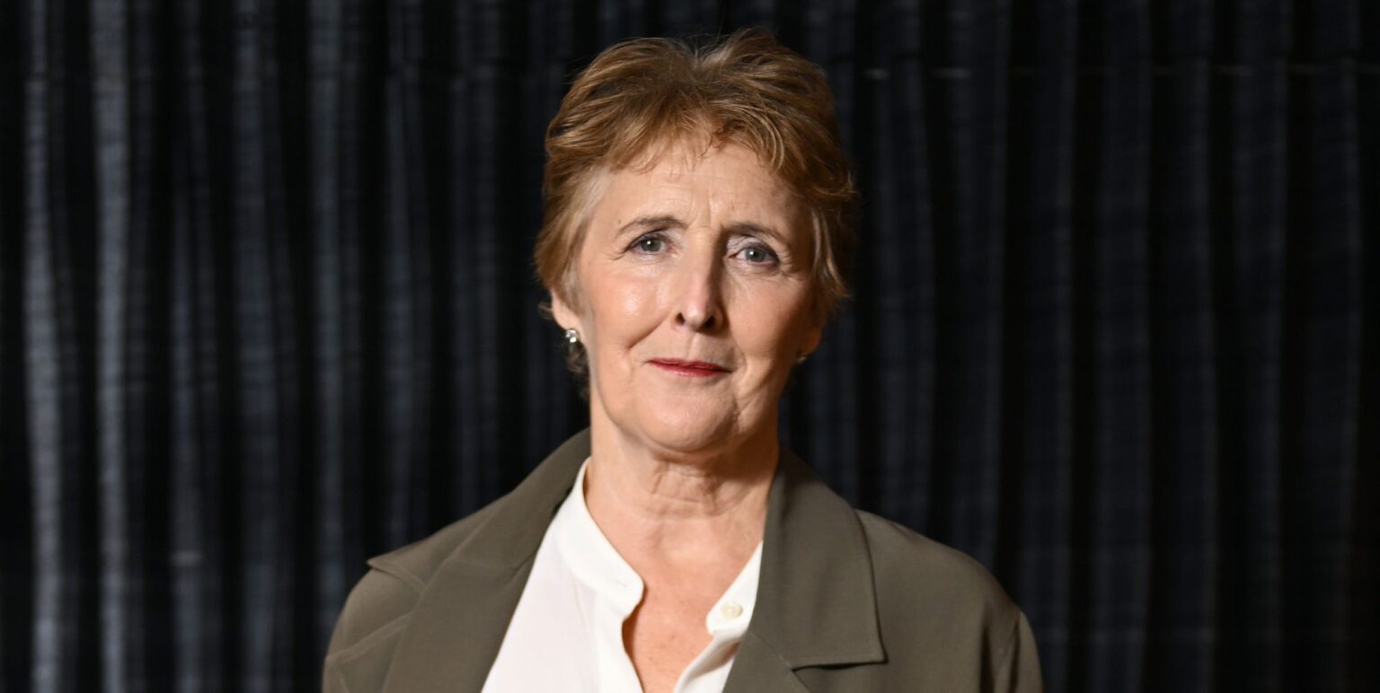 Fiona Shaw in a grey jacket, white shirt, against a black background.