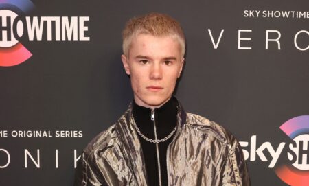 Young Royals star Edvin Ryding in a silver jacket, black jumper and necklace stood against a grey background.