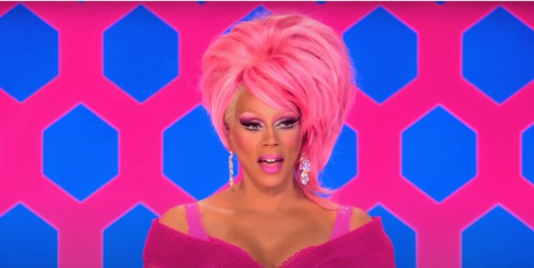 RuPaul on the Drag Rac judging panel in a pink outfit and pink wig with her mouth open.