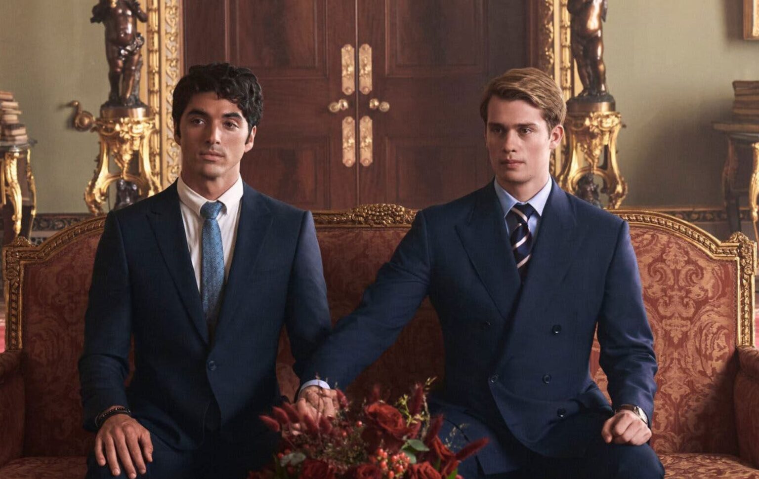 Red, White & Royal Blue still: Alex (Perez) and Prince Henry (Galitzine) sat on a royal red and gold sofa, both wearing suits. Henry has stretched his arm to hold Alex