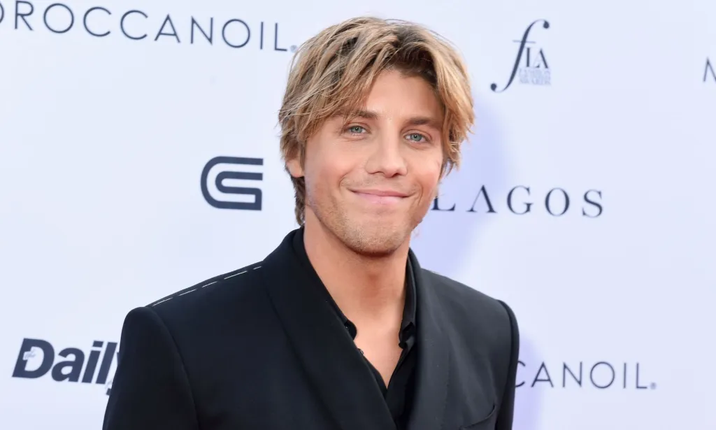 Lukas Gage dans un costume noir et une chemise avec des cheveux blonds séparés, souriant à la caméra.