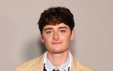 Noah Schnapp in a stripped white and blue shirt with a light beige jacket. Standing against a grey wall