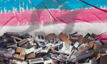 A pile of broken tech edited in front of a trans flag with the Grok logo superimposed over it.