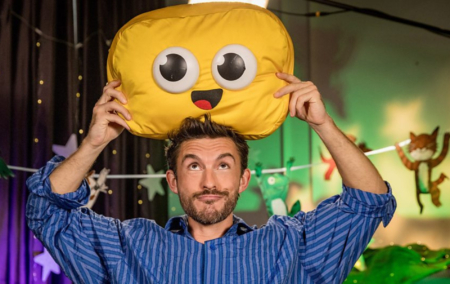 Jonathan Bailey holding a cuddly emoji smile cushion above his head