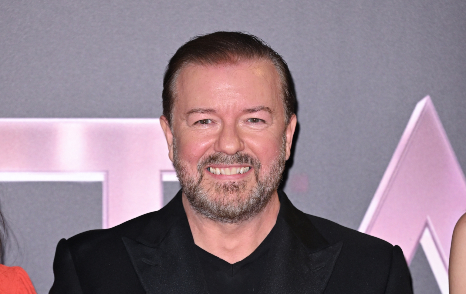 Ricky Gervais s'oppose aux critiques sur les blagues anti-trans : « Je pense que j'ai raison » Ricky Gervais in all black suit smiling at the camera
